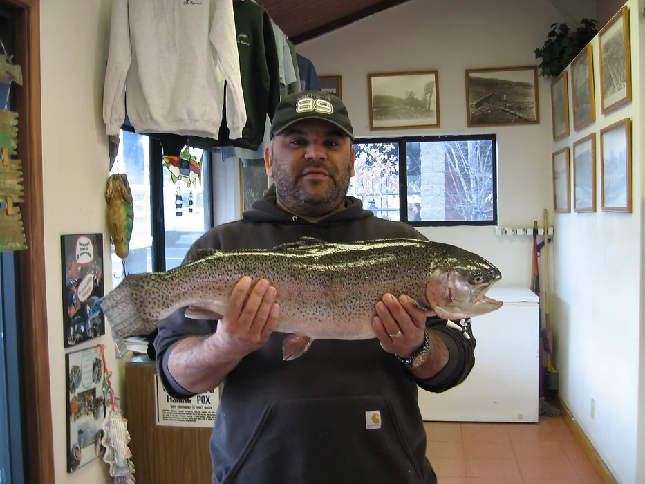 A person holding a large fish indoors, with a cap and hoodie on, surrounded by framed pictures and clothes hanging above.