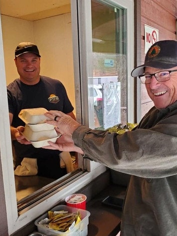 Two men are exchanging takeout boxes through a window, both smiling, with condiment packets on a counter.