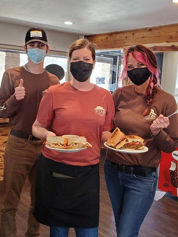 Three people wearing masks and holding plates of sandwiches in a restaurant setting.