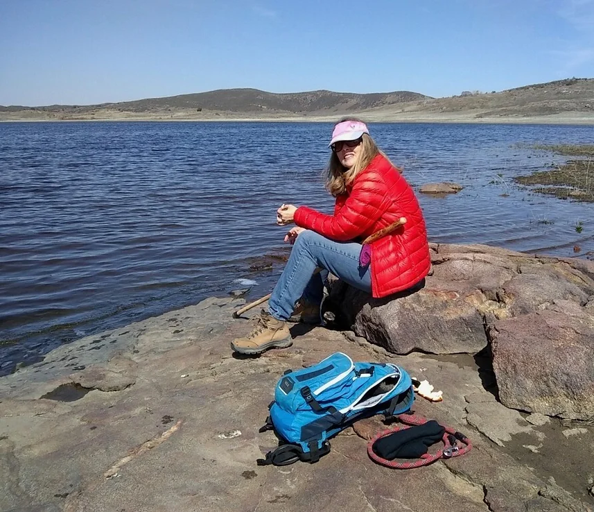 Person in red jacket sitting by a lake with a blue backpack on the rocks.