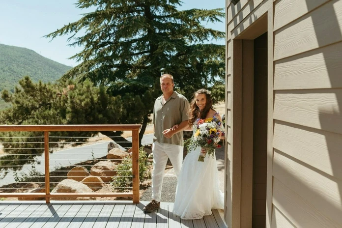 A bride holding a bouquet walks with a man on a deck outside, with trees and hills in the background.