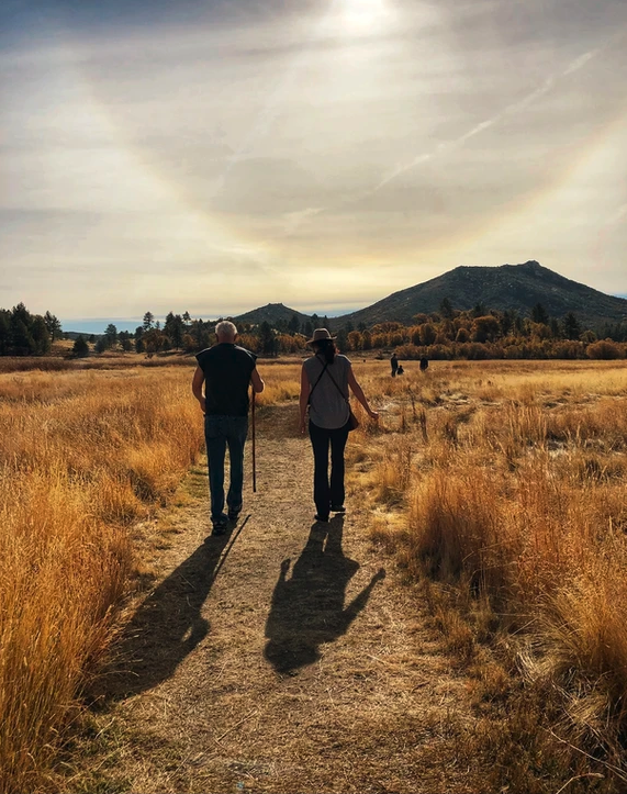 Two people walking on a path through a grassy field towards distant hills under a partly cloudy sky.