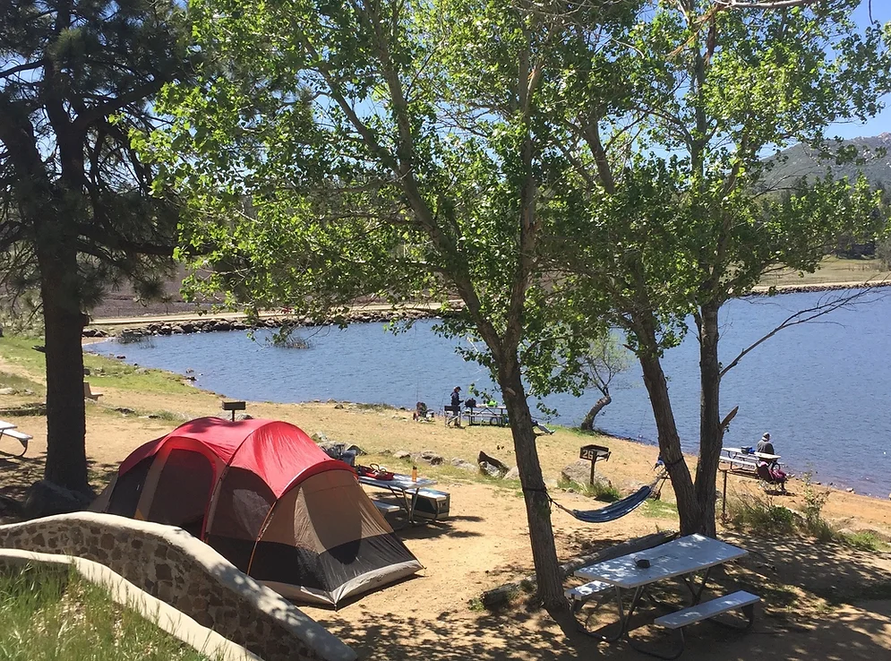 A lakeside campsite with a tent, hammock, picnic table, and trees, overlooking people relaxing by the water.