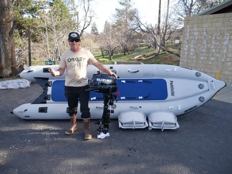 A person standing next to an inflatable boat with a Suzuki outboard motor, holding a drink, and wearing a cap and boots.