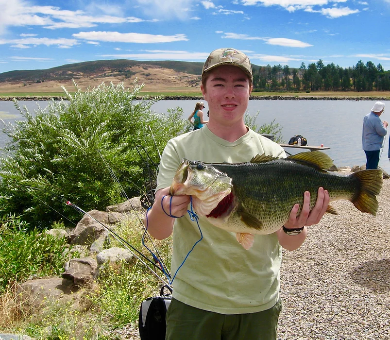 Person holding a large fish by a lakeside with fishing rods and another person fishing in the background.