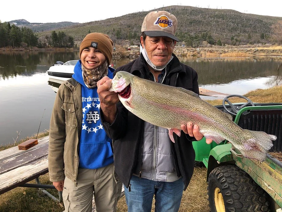 Two people smiling, holding a large fish by a lake with mountains and a boat in the background.