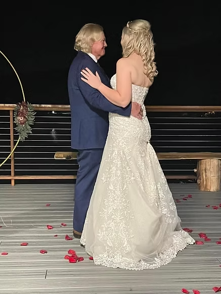 A couple dances at night; she's in a wedding dress, he's in a suit. Red petals are scattered on the wooden floor.