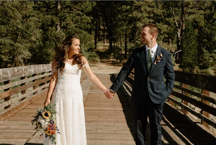 A bride and groom hold hands while smiling at each other on a wooden bridge, surrounded by trees.