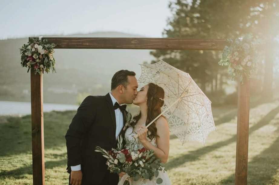 A couple kissing under a decorated arch outdoors, with the woman holding a parasol and bouquet.