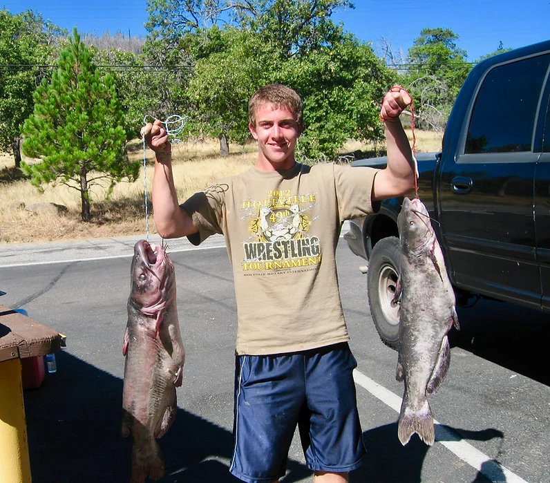 A person holding two large fish outdoors near a vehicle.