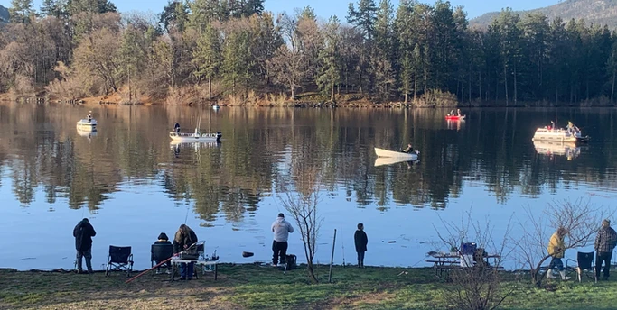 People fishing by a calm lake with boats, surrounded by trees and a clear sky.