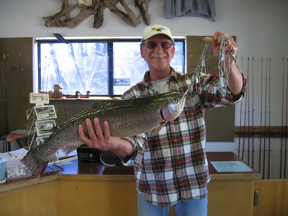 A man in a plaid shirt holds a large fish inside a shop with fishing gear and postcards.
