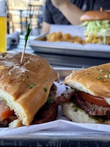 Two sandwiches with meat and tomatoes, fries, a burger, and a beer in the background on a table.