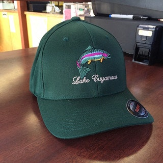 A green cap with "Lake Cuyamaca" and a colorful fish embroidered on it, placed on a wooden surface.