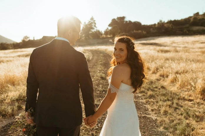 A couple in wedding attire holding hands, standing on a sunlit path in a field.