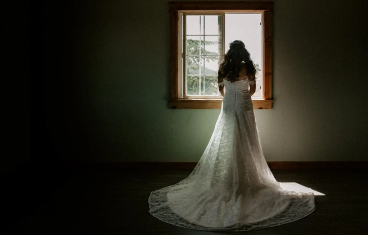 A bride in a white dress, standing in front of a window, facing outside.