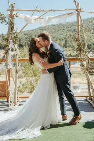A bride and groom kiss at an outdoor wedding ceremony under a rustic arch, surrounded by nature.