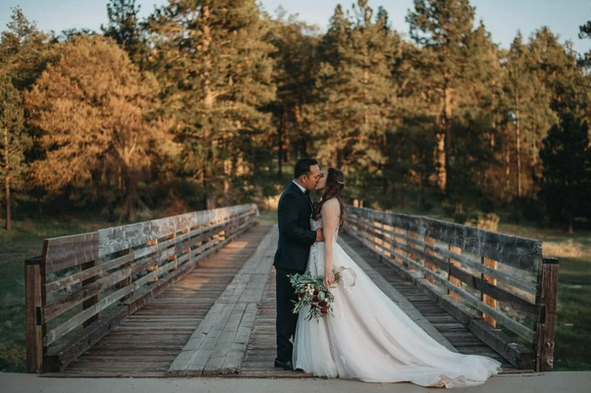 A couple in wedding attire is kissing on a wooden bridge surrounded by trees.