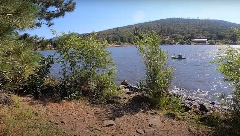 A scenic lake with trees and shrubs in the foreground, a kayaker on the water, and a hill with trees in the background.