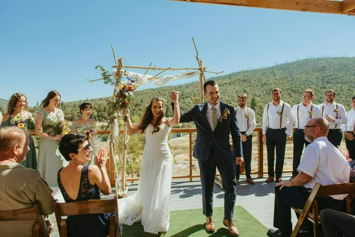 A wedding ceremony outdoors, with a couple under an arch, surrounded by friends and family applauding.