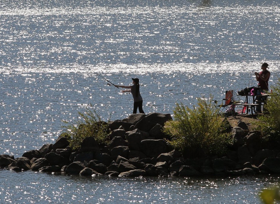 Two people fishing by a sparkling lake, one casting a line, the other preparing equipment, surrounded by rocks and bushes.