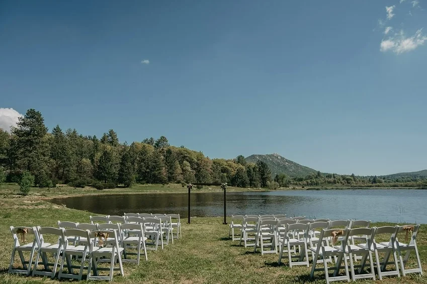 Outdoor wedding setup by a lake, with white chairs facing an arch, surrounded by trees and mountains under a clear blue sky.
