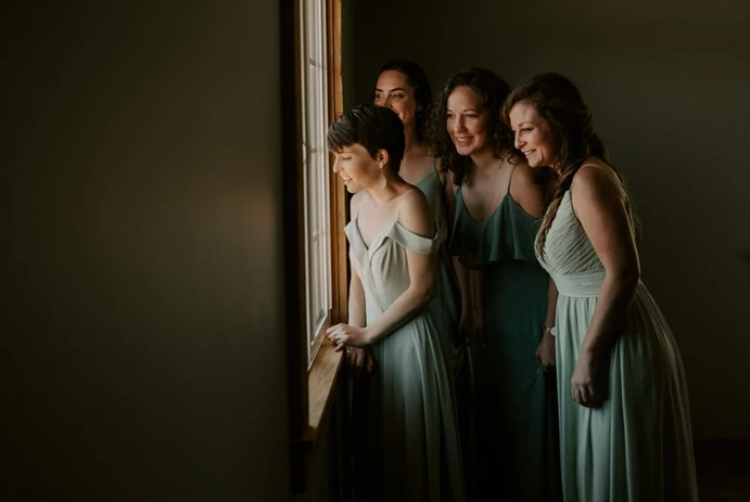 Four women in dresses eagerly look out a window, smiling in anticipation.