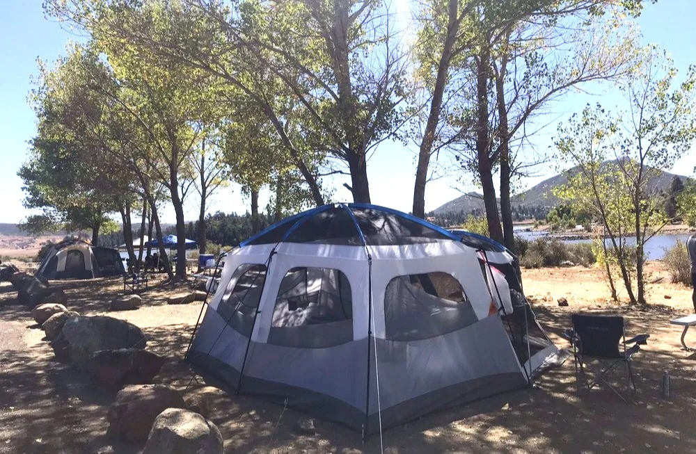Tents set up among trees by a lake with mountains in the background, and camping chairs nearby.