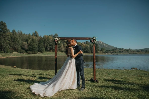 A couple kissing under a wooden arch by a lake, surrounded by nature, during a wedding ceremony.
