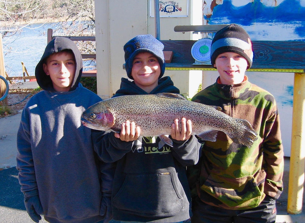 Three kids in hoodies, smiling and holding a large fish outdoors.