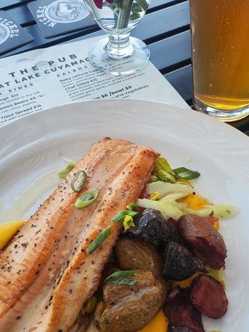 A plate with grilled fish, roasted potatoes, and vegetables, alongside a glass of beer and a menu.