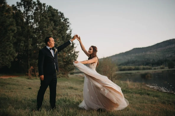 A couple dances outdoors, dressed in wedding attire, surrounded by nature with trees and water in the background.