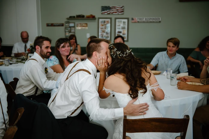 A couple wearing wedding attire kisses at a reception table with guests around them.