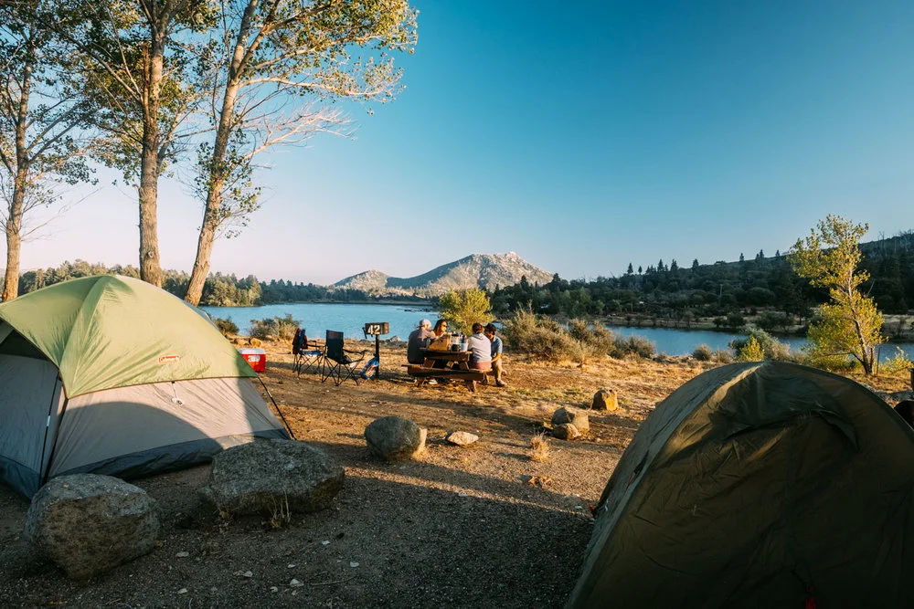 Camping site by a lake with tents, people at a picnic table, trees, and mountains in the background.