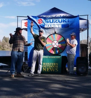 Three people stand in front of a spinning wheel under a Suzuki Marine tent with a "Western Outdoor News" sign.