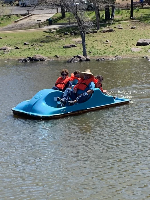 Four people in life vests on a blue paddle boat on a lake.