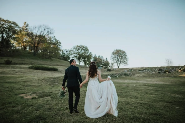 A couple in wedding attire walks through a grassy field, the groom holding flowers, with trees in the background.