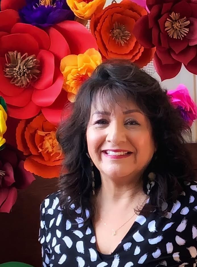 A smiling woman in front of vibrant, colorful paper flowers, wearing a black patterned top with earrings. Joyful ambiance!