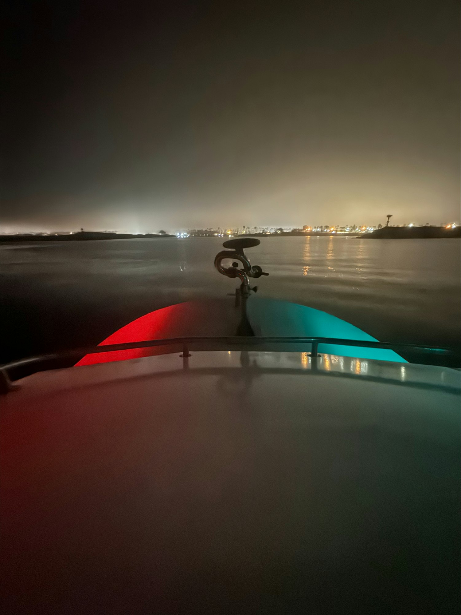 Nighttime boat view with red and green lights reflecting on water, distant city lights visible on the horizon.