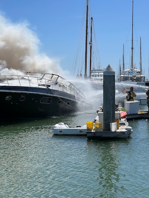 A boat on fire with heavy smoke, firefighter hosing water, at a marina.