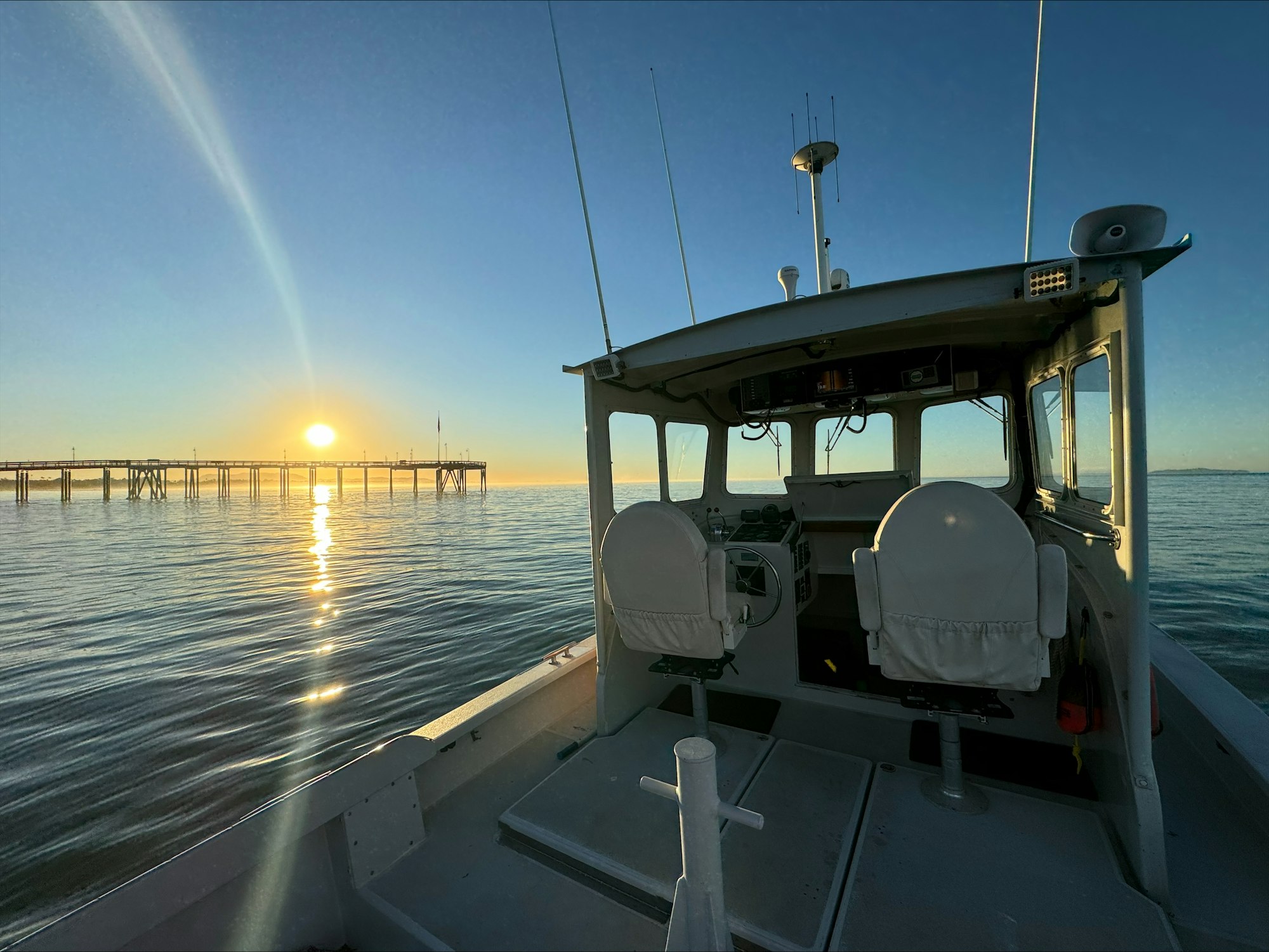 Boat interior facing the sea with a pier and a sunset on the horizon.