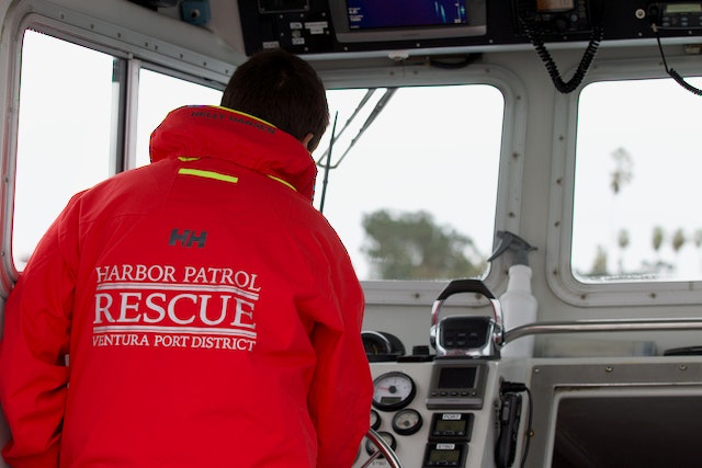 A person in a red "Harbor Patrol Rescue" jacket inside a boat's cockpit with navigational equipment.
