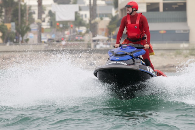 A person in a red life vest riding a jet ski on water, with splashes around and buildings in the background.