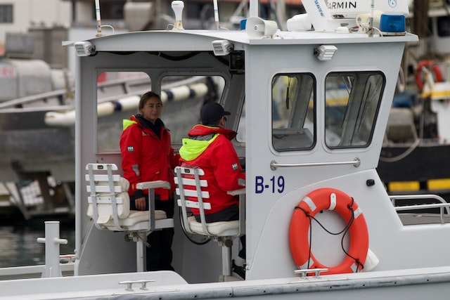 Two people on a boat with "B-19" visible, wearing safety gear, and a lifebuoy attached nearby.