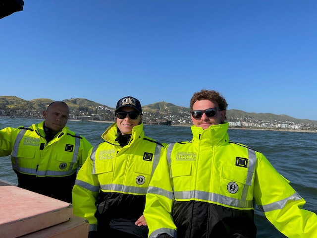 Three people in bright yellow rescue jackets on a boat with hills and water in the background.