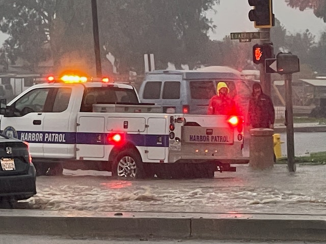A Harbor Patrol vehicle and people in a flooded street, emergency lights flashing, during heavy rain.