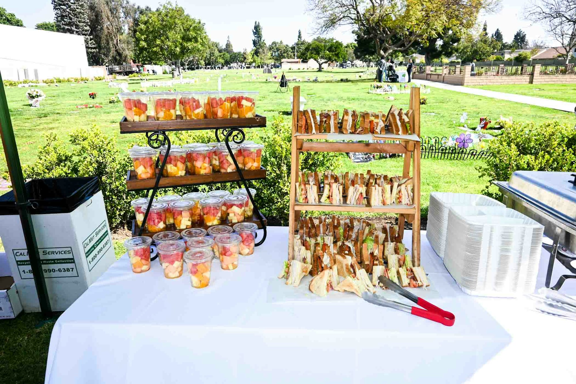 Sandwiches and fruit cups on a table outdoors with plates and a trash bin nearby.