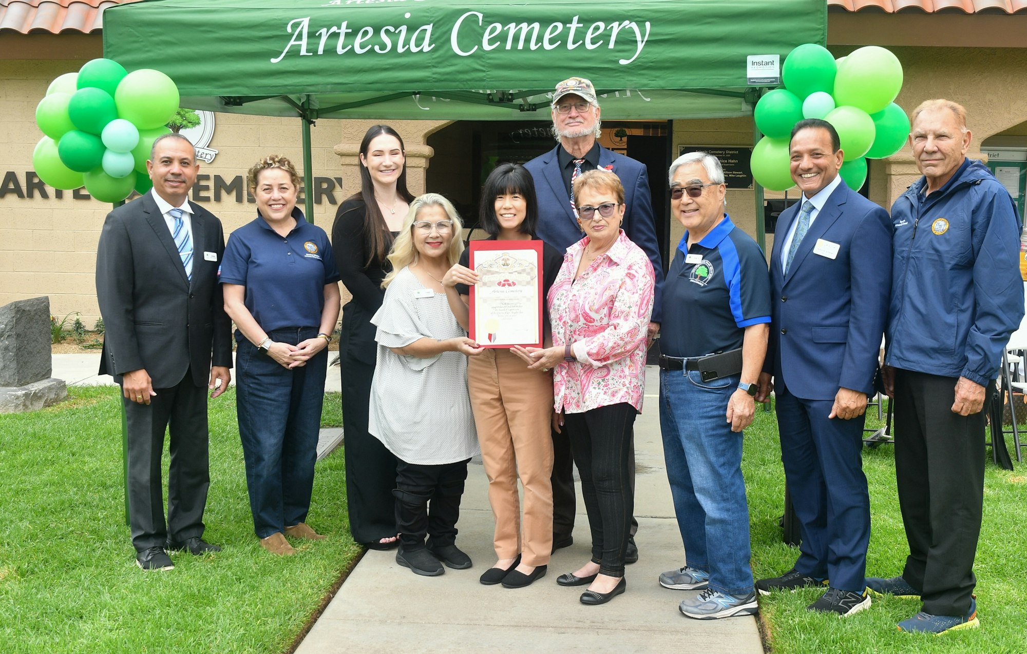 Group of people at an event in front of "Artesia Cemetery" tent, with balloons and a woman holding a framed certificate.