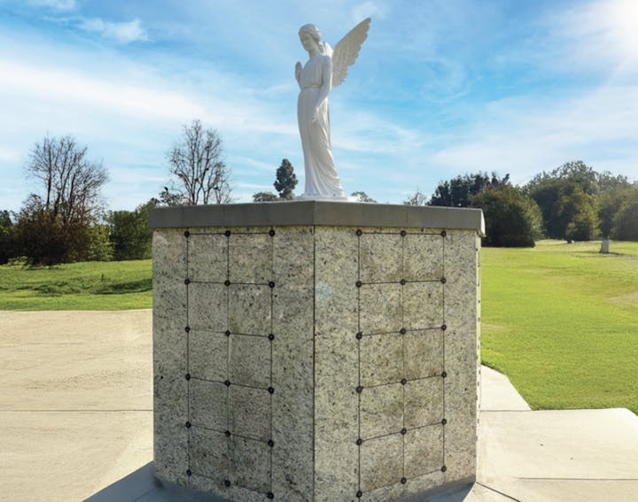 An angel statue atop a stone structure in a cemetery, surrounded by grass and trees under a blue sky.