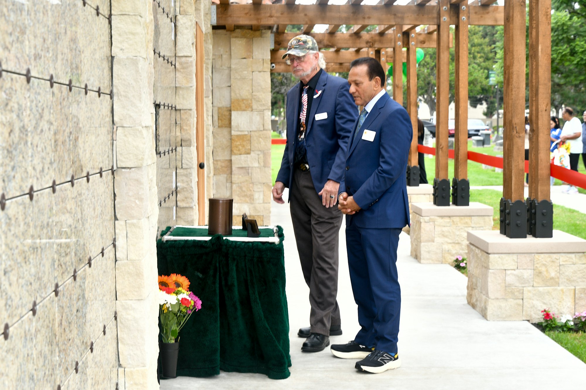 Two men in formal attire are standing next to a table with flowers during a daytime outdoor event.